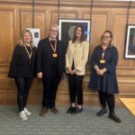 Four people wearing orange lanyards stand side by side in a wood‑panelled room, with framed circular artworks displayed on the wall behind them.