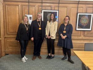 Four people wearing orange lanyards stand side by side in a wood‑panelled room, with framed circular artworks displayed on the wall behind them.